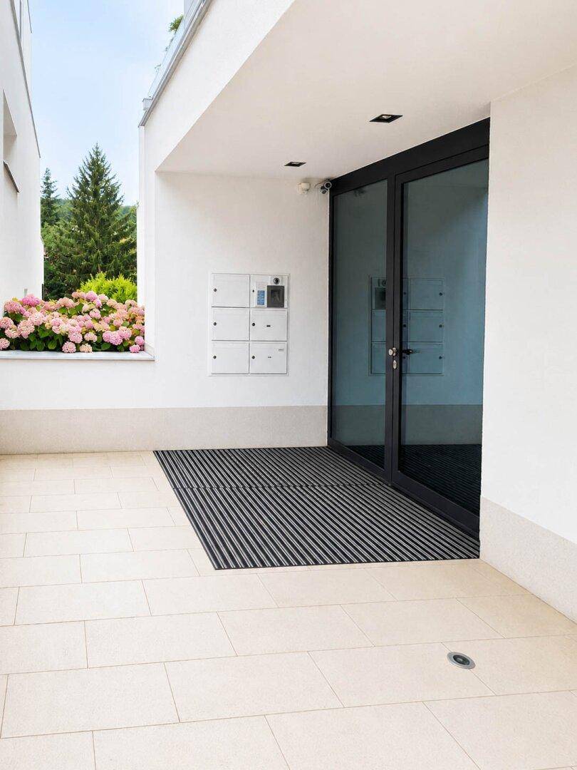 Eingangsbereich eines modernen Gebäudes mit großen Glastüren und Gegensprechanlage.