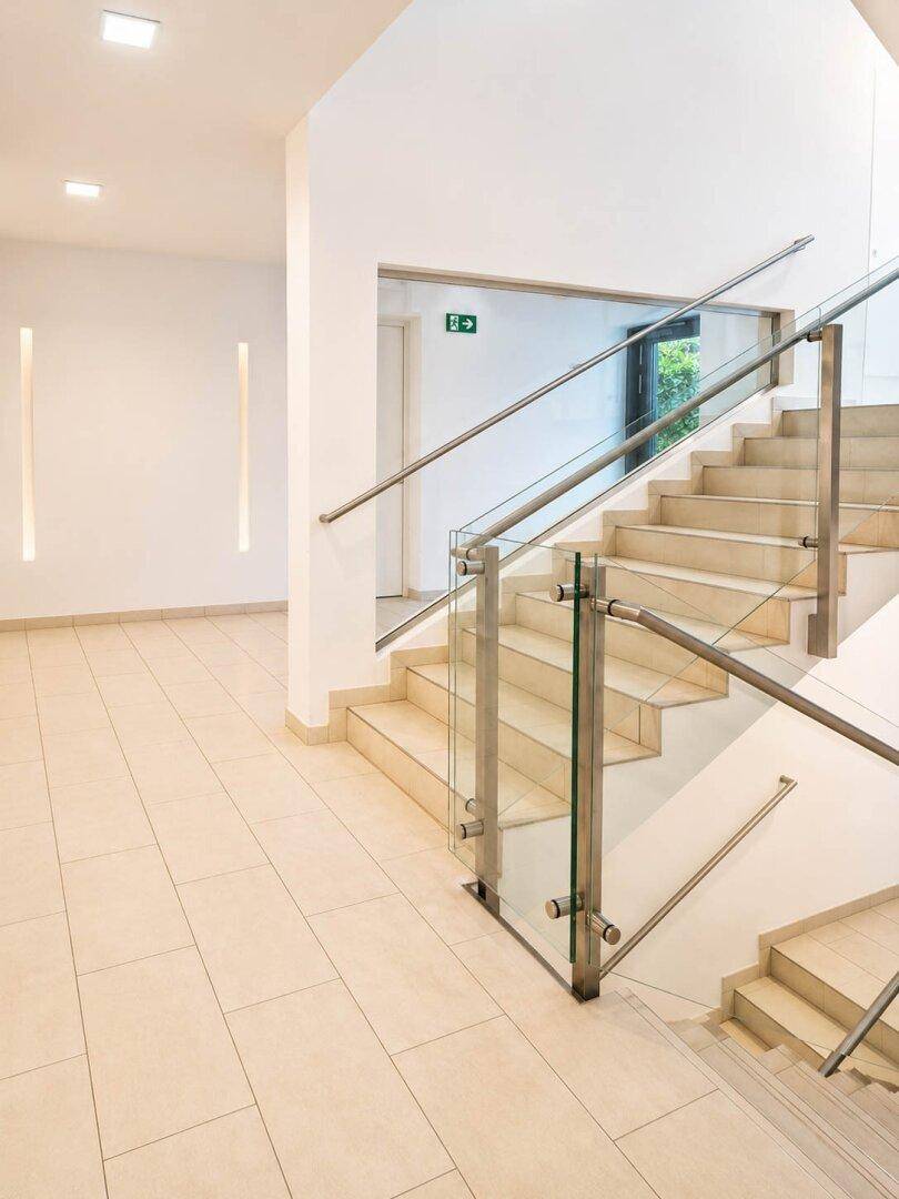 Heller Treppenaufgang mit Glasgeländer und Fliesenboden in einem modernen Gebäude.