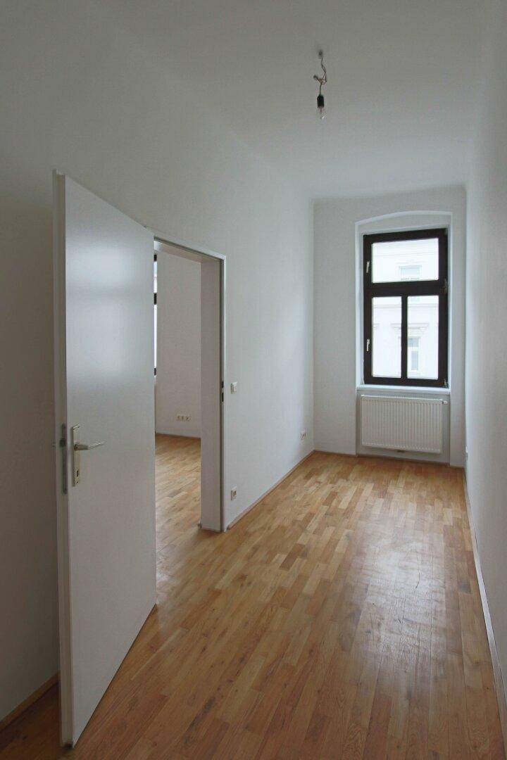 Leerer Raum mit Holzboden und einem Fenster, der als Flur oder Durchgangszimmer dienen könnte.