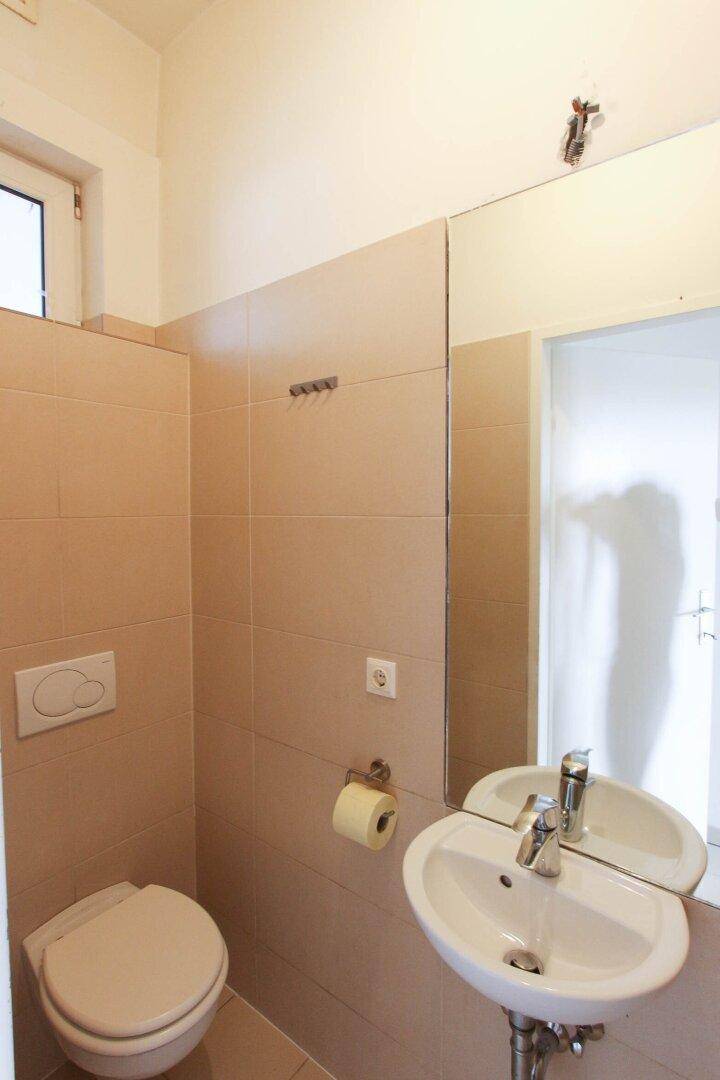 Kleines, funktionales WC mit beigefarbenen Fliesen, Toilette und einem einzelnen Waschbecken.