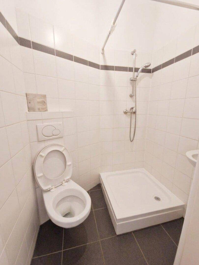 Kleines Badezimmer mit Duschbereich, wandhängendem WC und weißen Fliesen an den Wänden.