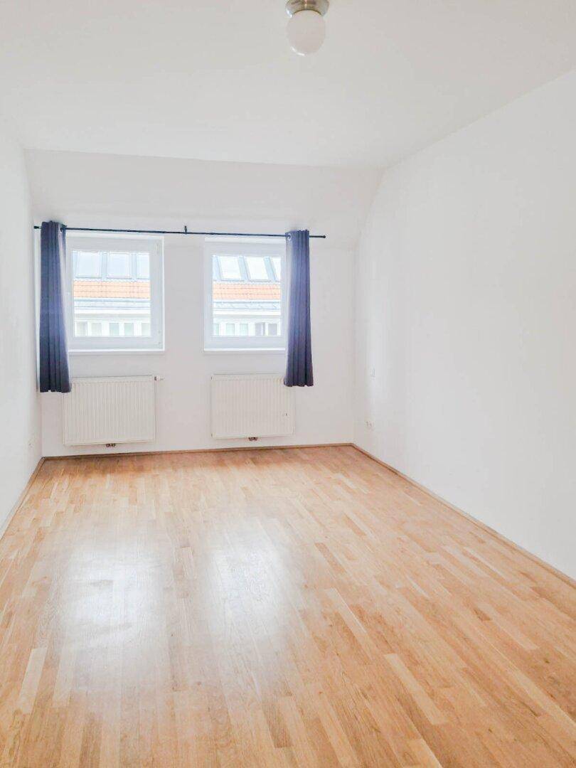 Helles Schlafzimmer mit Parkettboden, zwei Fenstern und blauen Vorhängen.