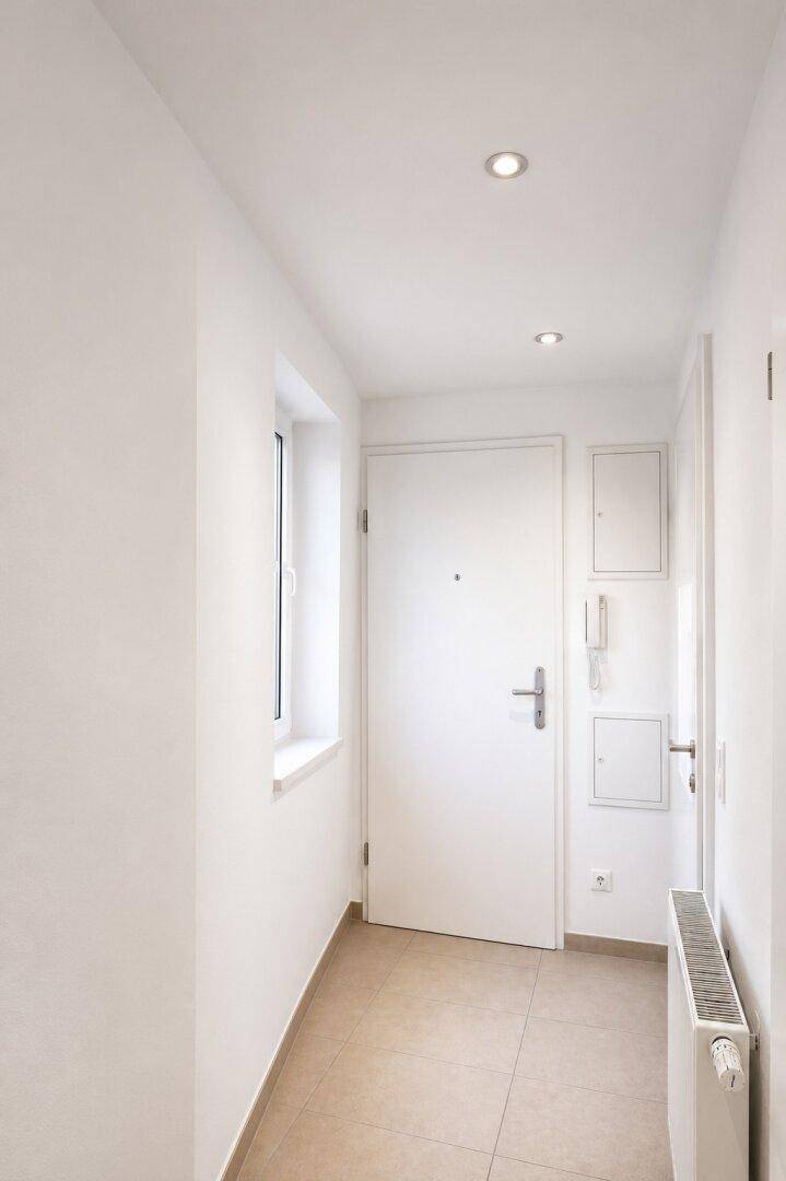Heller Eingangsbereich mit Fliesenboden, weißer Tür und integrierten Deckenleuchten.