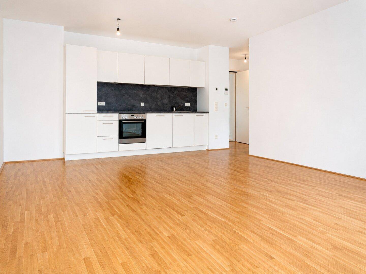 Offener Wohnbereich mit moderner weißer Einbauküche, dunkler Rückwand und hellem Parkettboden.