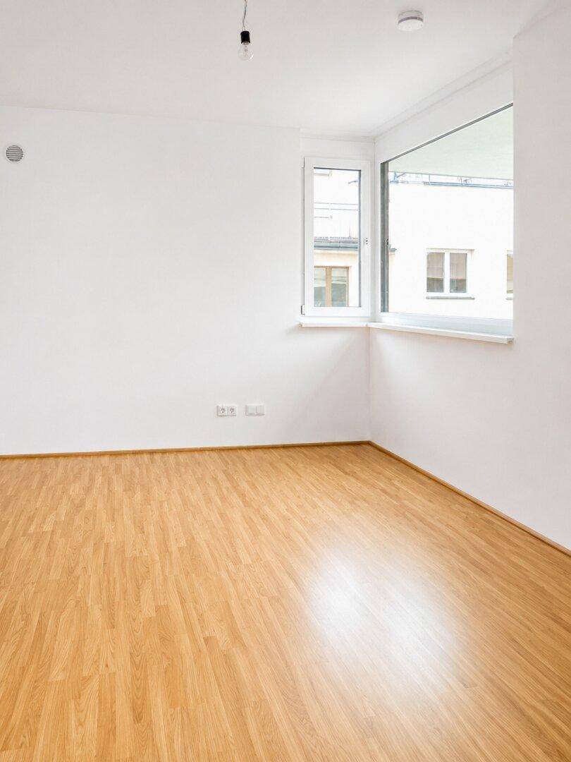 Heller Raum mit Parkettboden, weißen Wänden und einem großen Eckfenster für viel Tageslicht.