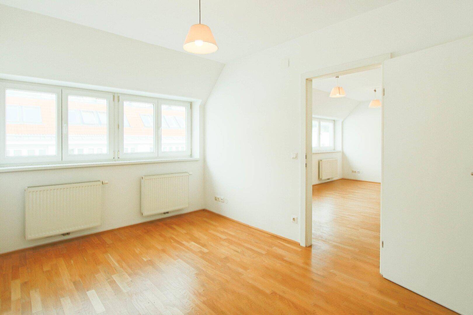 Helles Schlafzimmer mit Parkettboden und mehreren Fenstern, verbunden mit einem weiteren Raum.