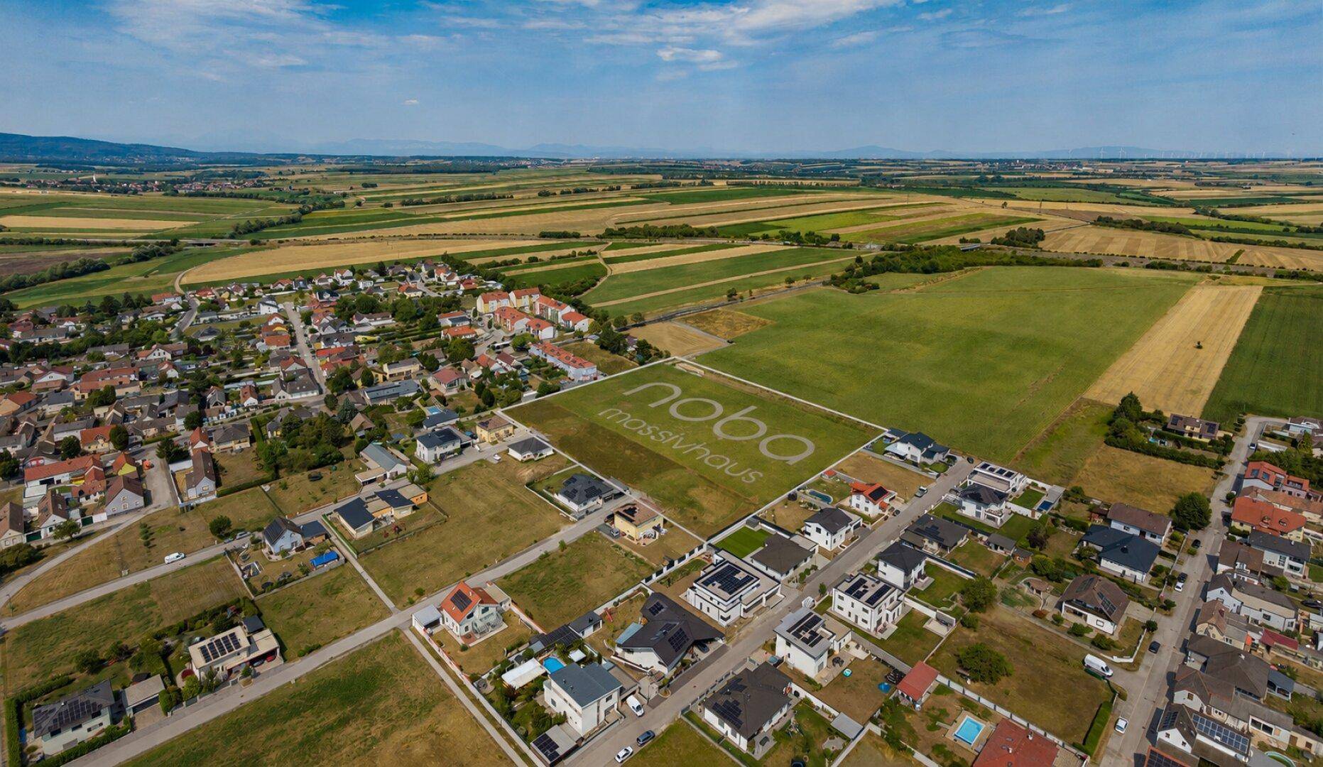 Weitläufige Luftansicht eines Baugrundstücks, umgeben von Wohnhäusern und landwirtschaftlichen Flächen.