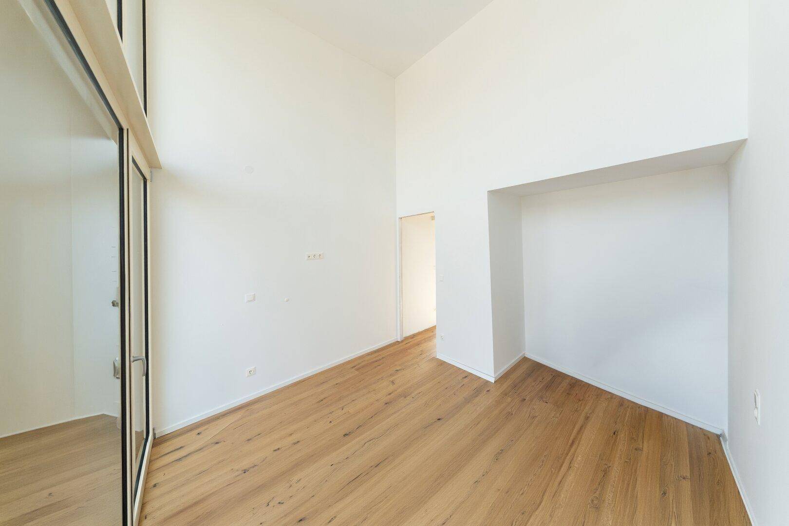 Geräumiger, heller Raum mit Holzboden, hohen Decken und einem Nischenbereich.