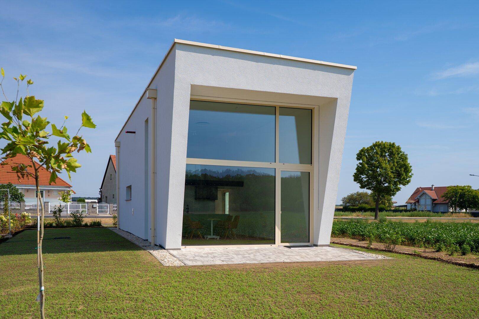 Moderne Hausfassade mit großen Fenstern und kleiner Terrasse, umgeben von Grünflächen.