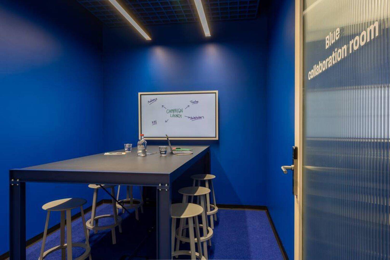 Lebhafter blauer Besprechungsraum mit einem Tisch, Hockern und einem Whiteboard für kreative Zusammenarbeit.