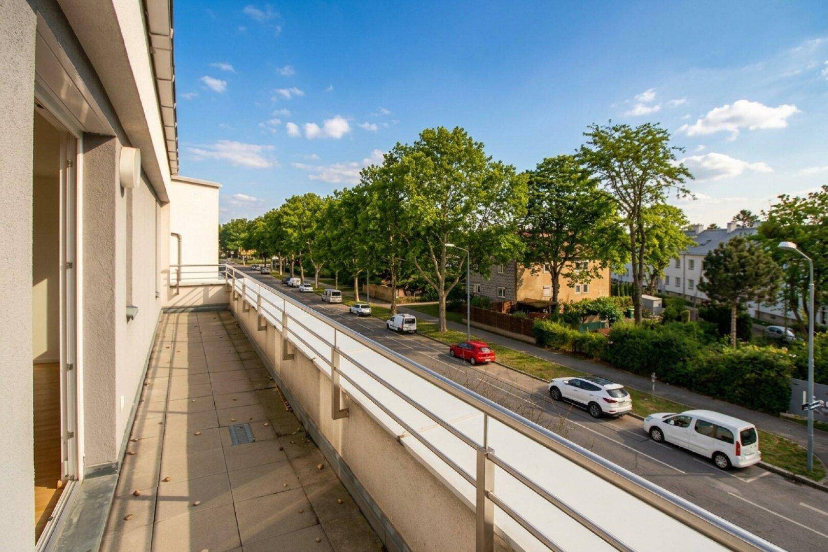 Langer Balkon mit Blick auf eine von Bäumen gesäumte Straße und vorbeifahrende Autos.