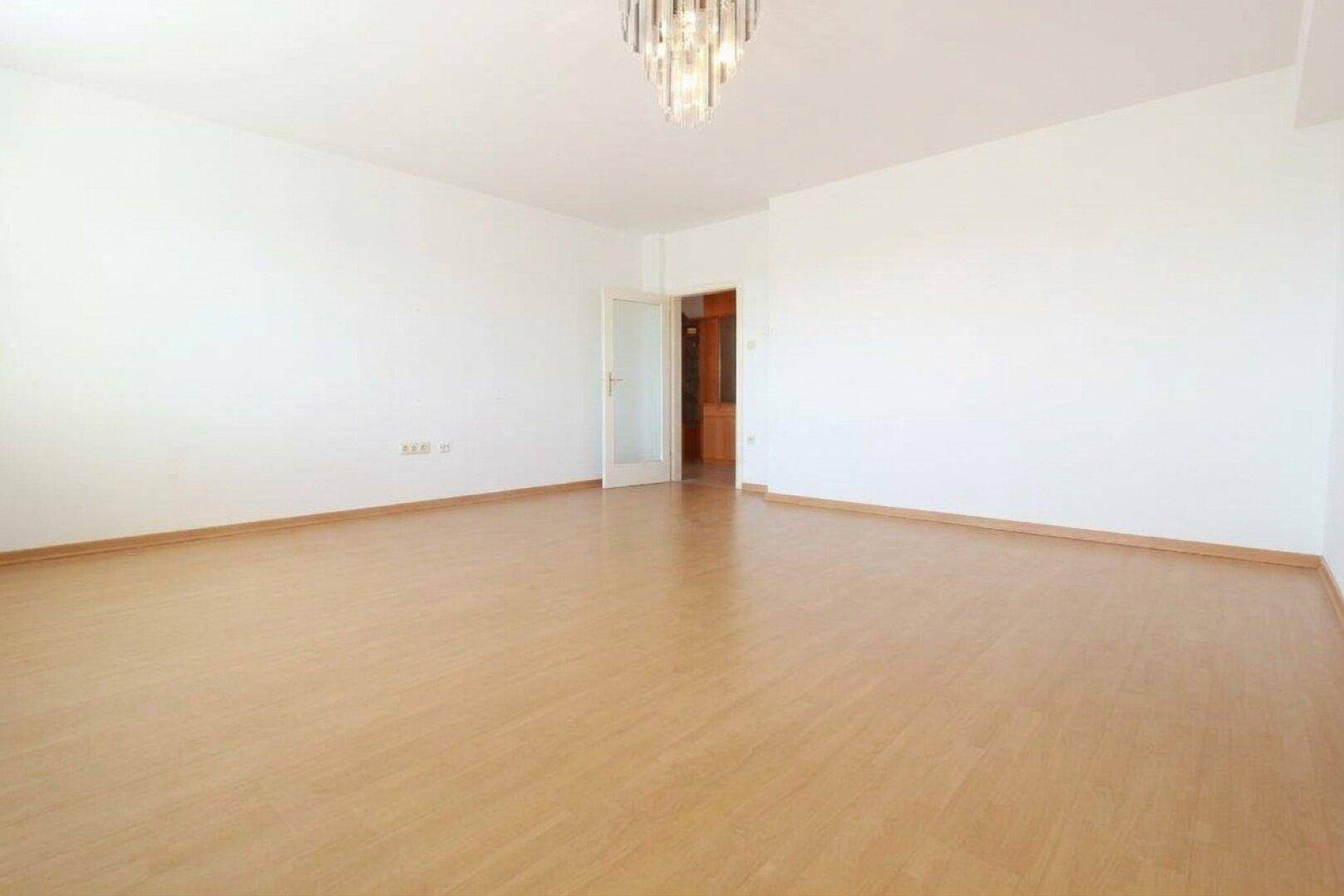 Geräumiges, unmöbliertes Wohnzimmer mit Laminatboden und einem eleganten Kronleuchter an der Decke.