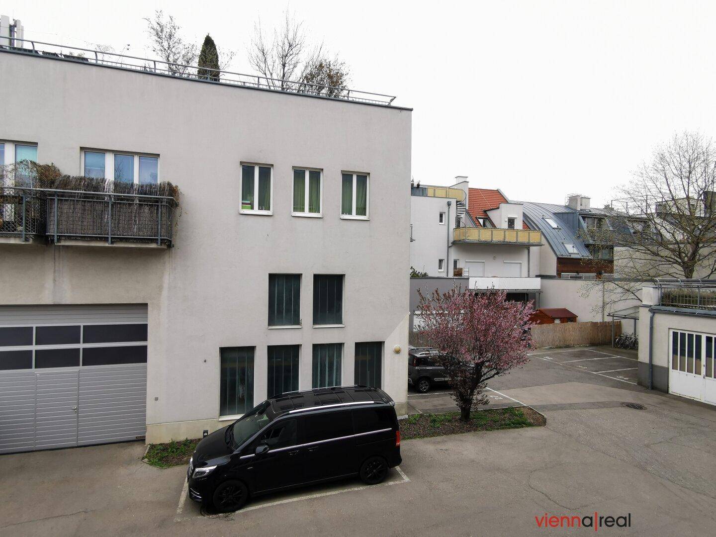 Außenansicht eines modernen Gebäudes mit Parkplätzen und einem kleinen Baum im Vordergrund, umgeben von weiterer Bebauung.