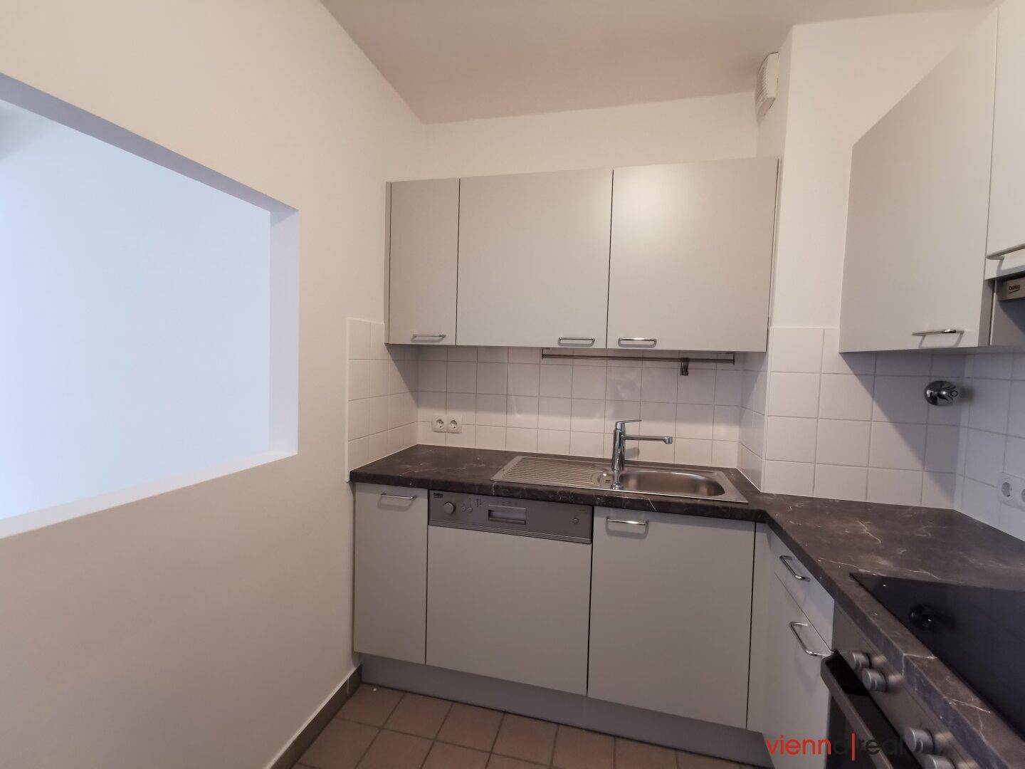 Kompakte Küche mit weißen Schränken, Spülbecken und Durchreiche zu einem angrenzenden Raum, auf Fliesenboden.