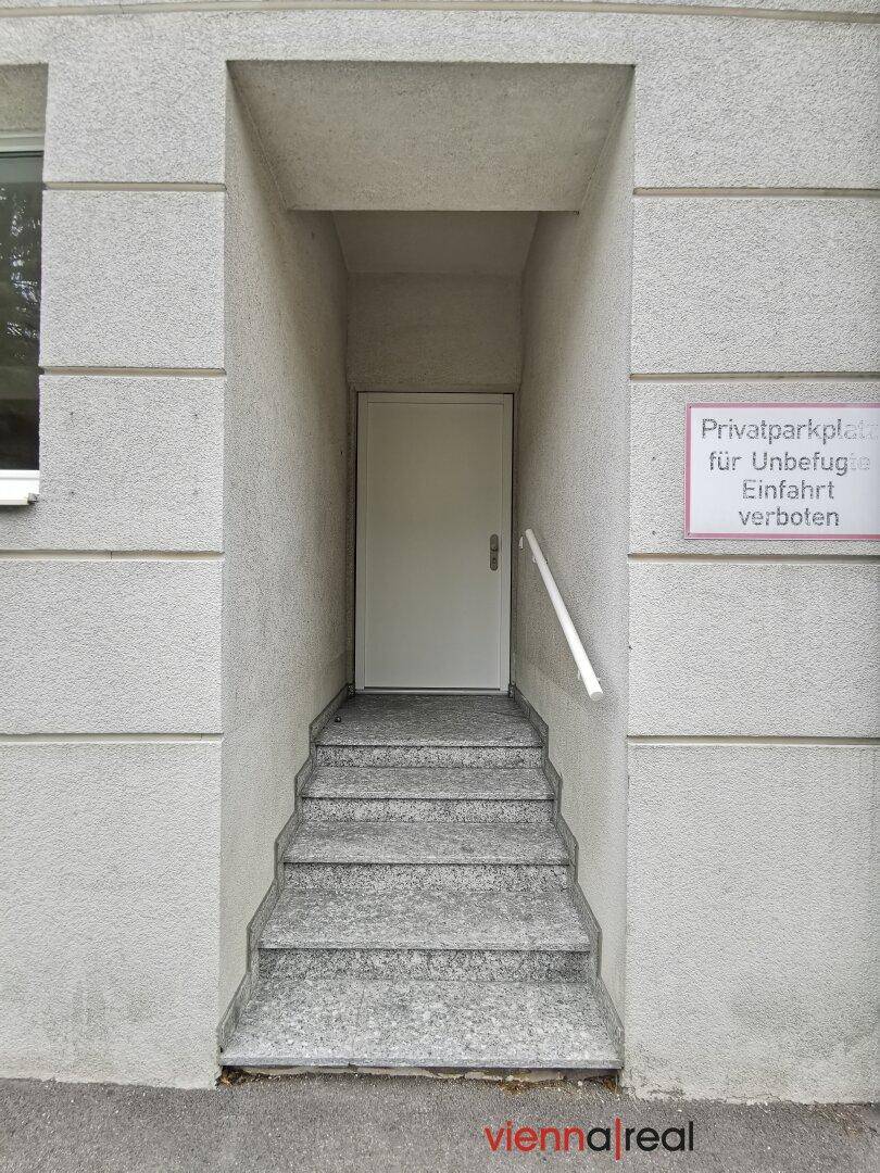 Eingangsbereich des Gebäudes mit Steintreppe und Handlauf, führt zu einer weißen Tür, umgeben von Putzfassade.