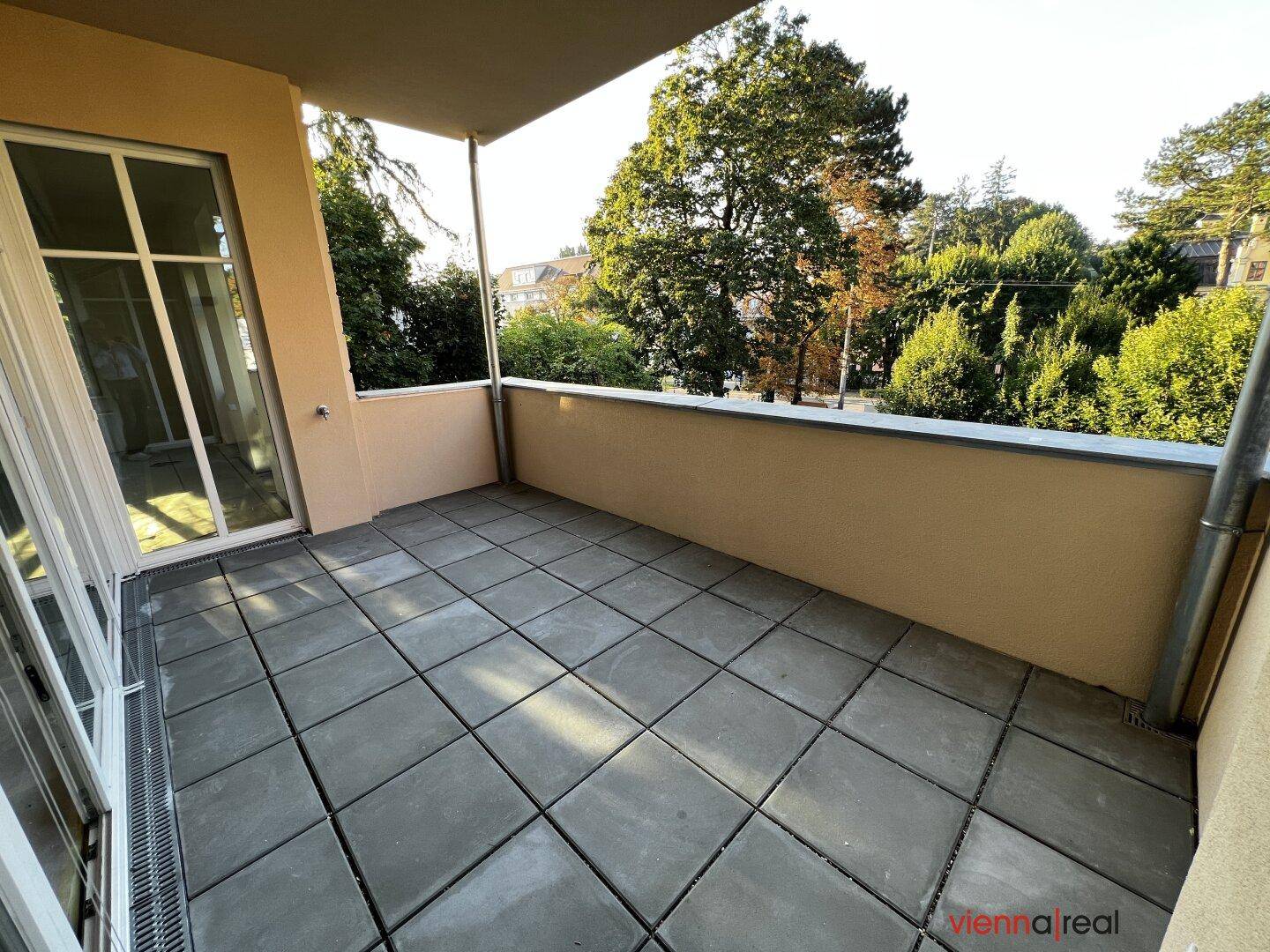 Heller Balkon mit Fliesenboden und Blick auf die umliegenden Bäume und Gebäude.