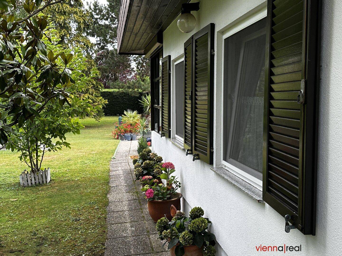 Seitenansicht des Hauses mit Fensterläden und einem gepflasterten Weg, umgeben von Grün.