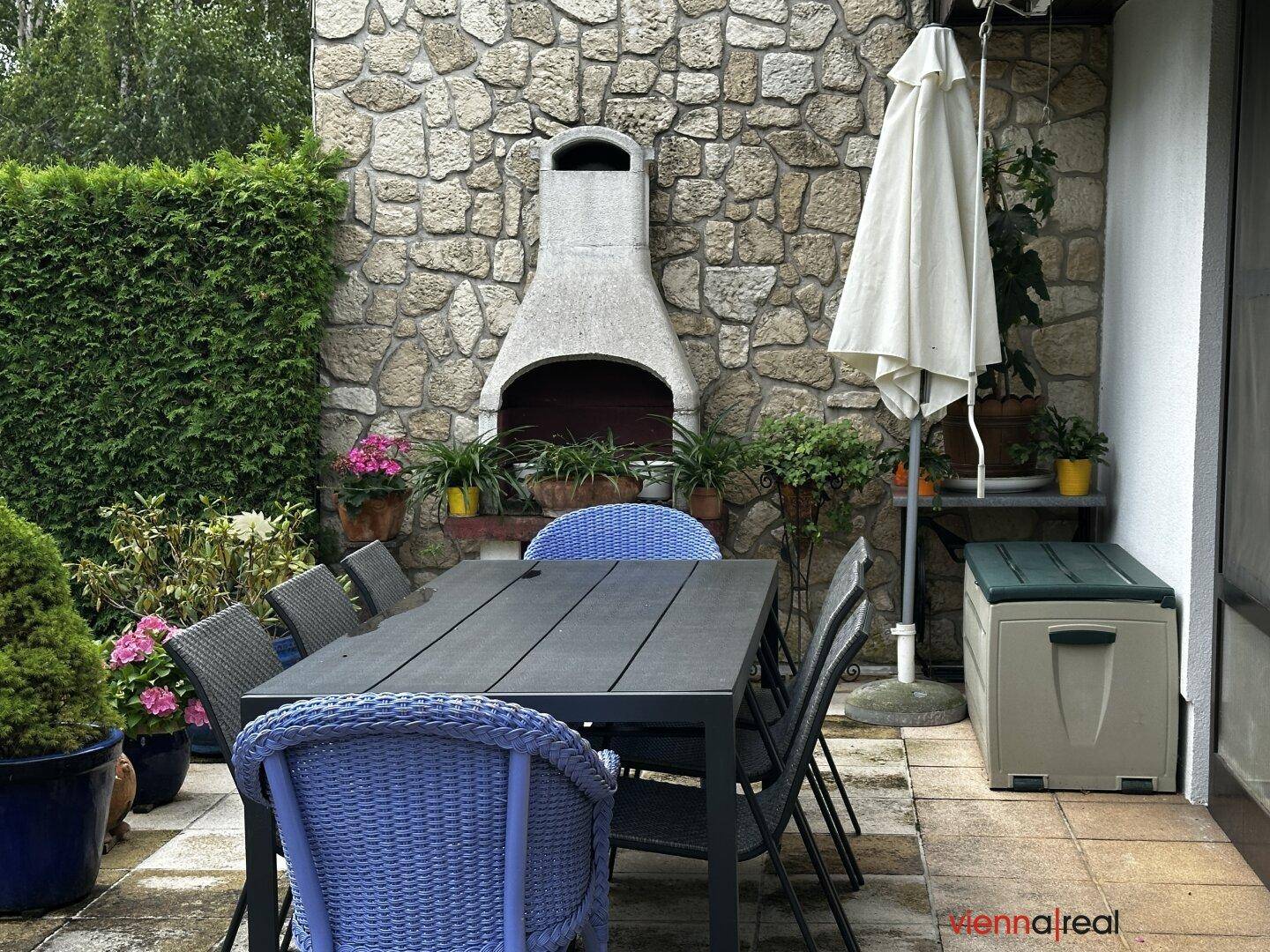 Detailansicht der Terrasse mit Steingrill, Esstisch und blauen Stühlen für Mahlzeiten im Freien.