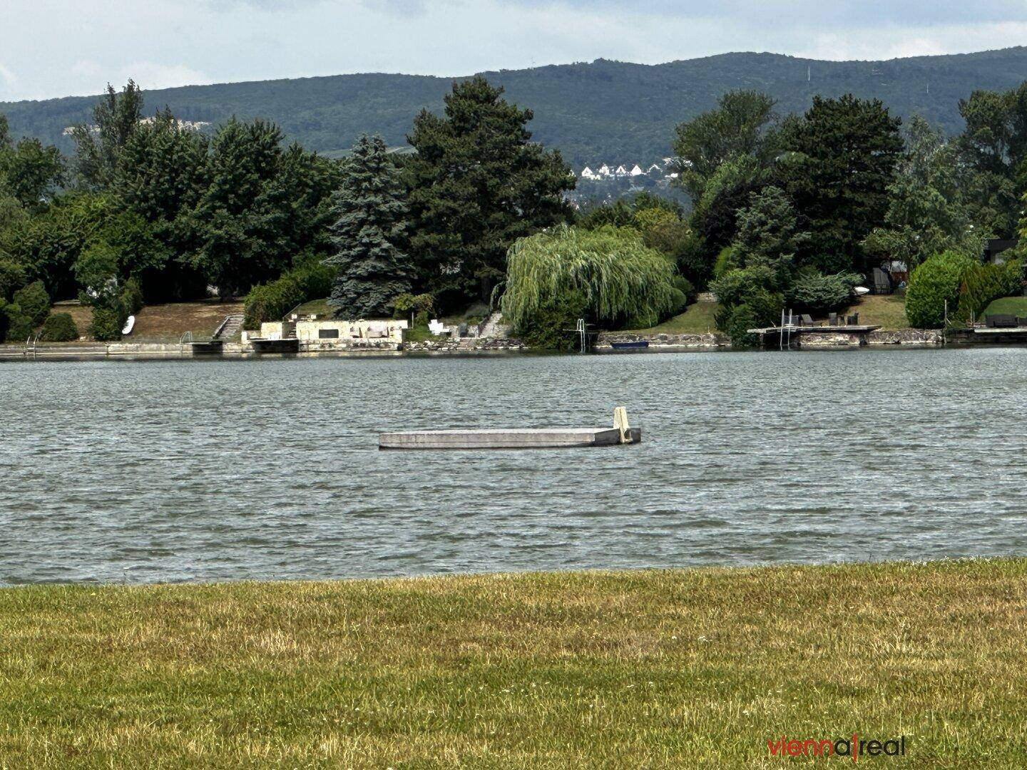 Weitläufiger Blick auf einen See mit bewaldeten Hügeln im Hintergrund unter bewölktem Himmel.