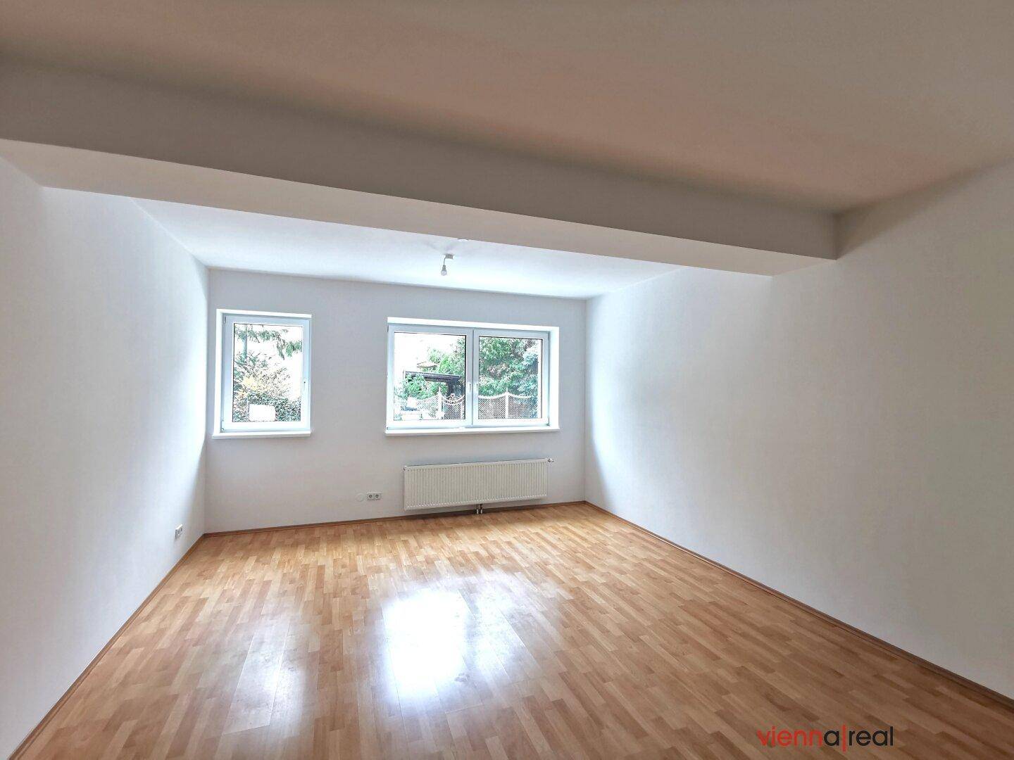 Geräumiger, unmöblierter Wohnbereich mit hellem Holzboden und zwei Fenstern, die den Raum erhellen.