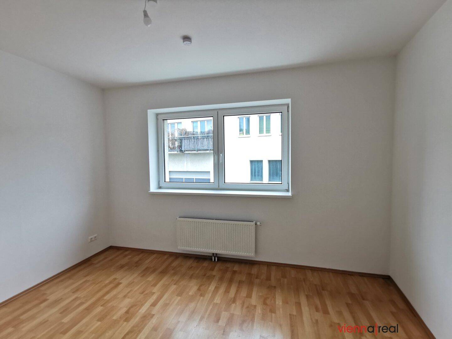 Unmöblierter Raum mit Holzboden, weiß gestrichenen Wänden und einem großen Fenster mit Blick nach draußen.