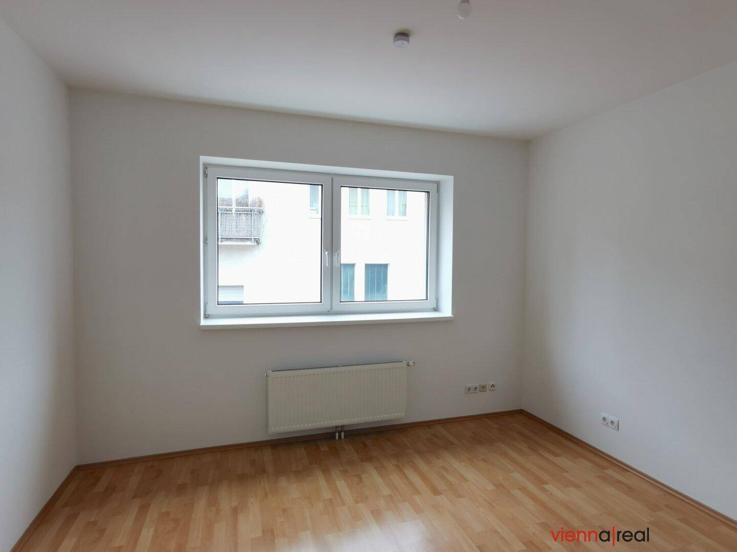 Heller, leerer Raum mit Holzboden und einem großen Fenster, das viel Tageslicht hereinlässt.