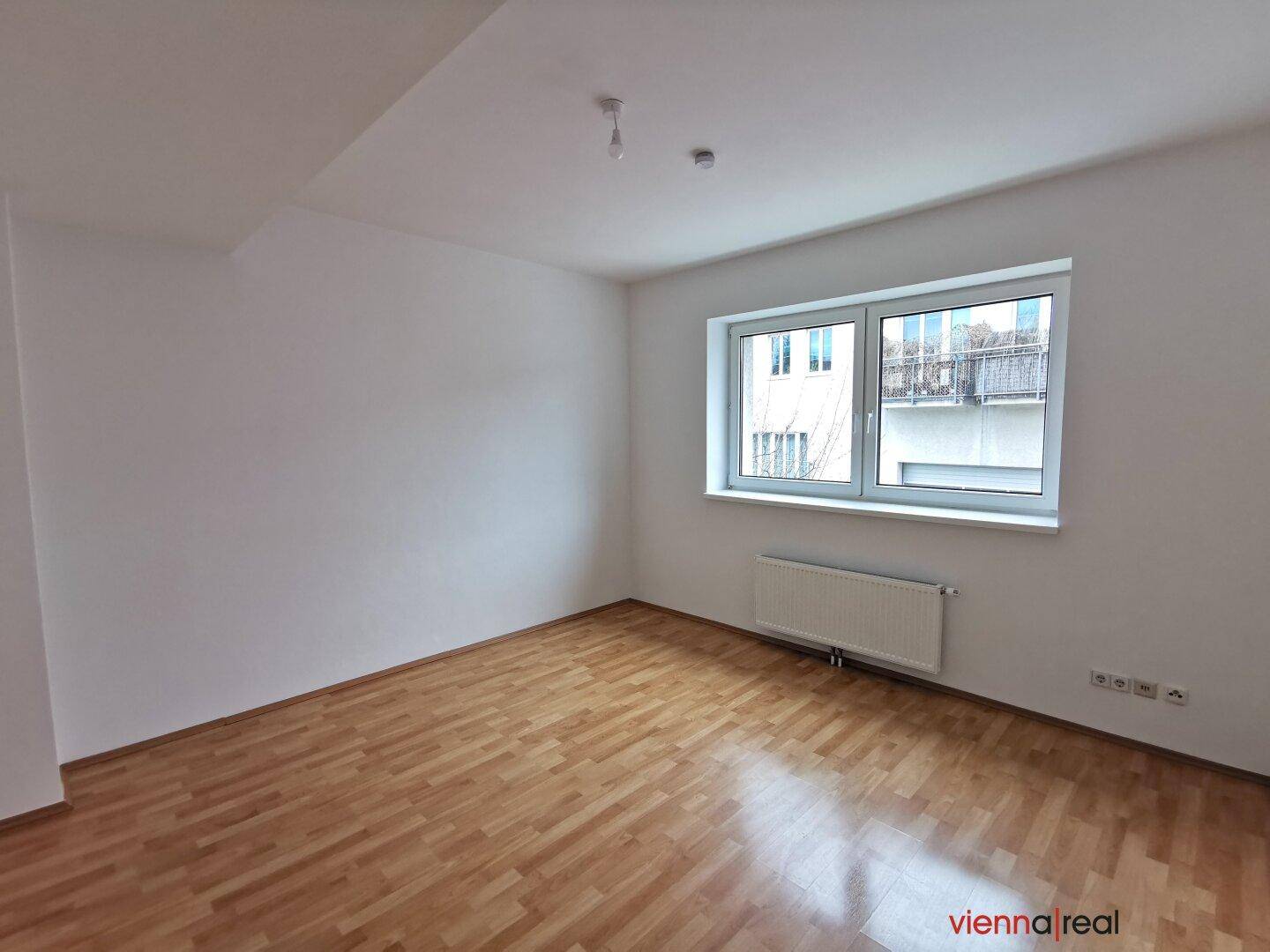 Geräumiger, unmöblierter Raum mit Holzboden und einem großen Fenster, das den Raum hell erscheinen lässt.