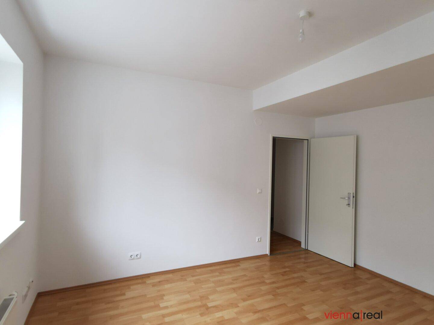 Heller Raum mit Holzboden und einer offenen Tür, die den Blick in einen weiteren Bereich freigibt.