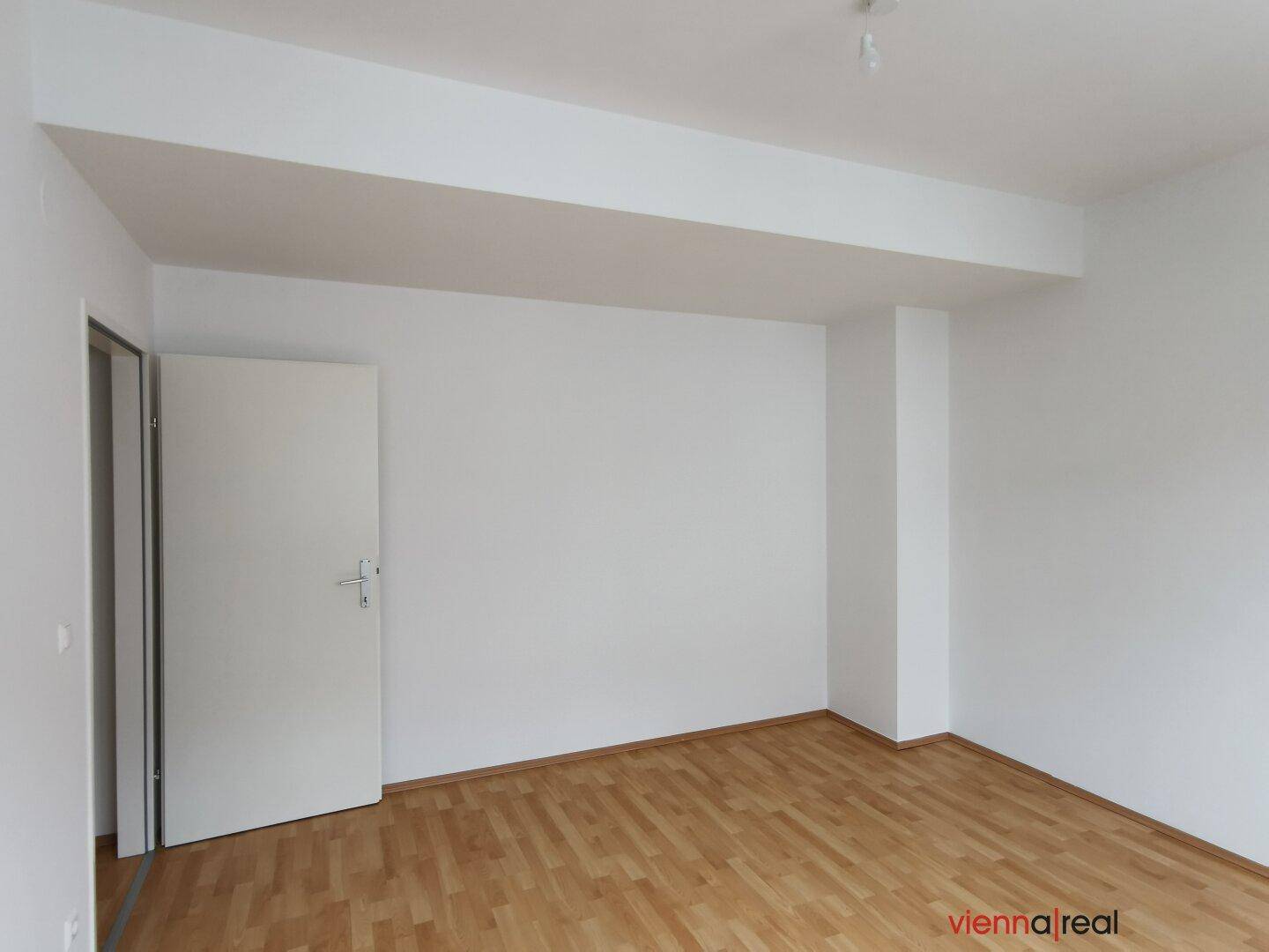 Unmöblierter Raum mit Holzboden, weißen Wänden und einer Tür, die zu einem anderen Zimmer führt.
