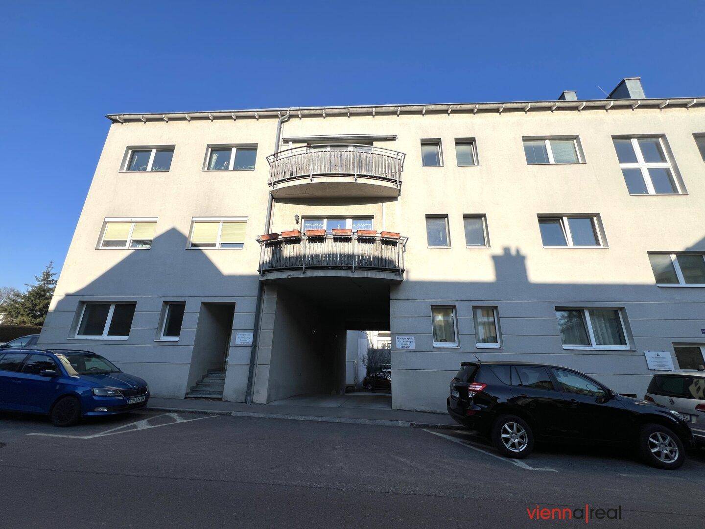 Mehrfamilienhaus mit Balkonen und einem Durchgang, umgeben von geparkten Autos.