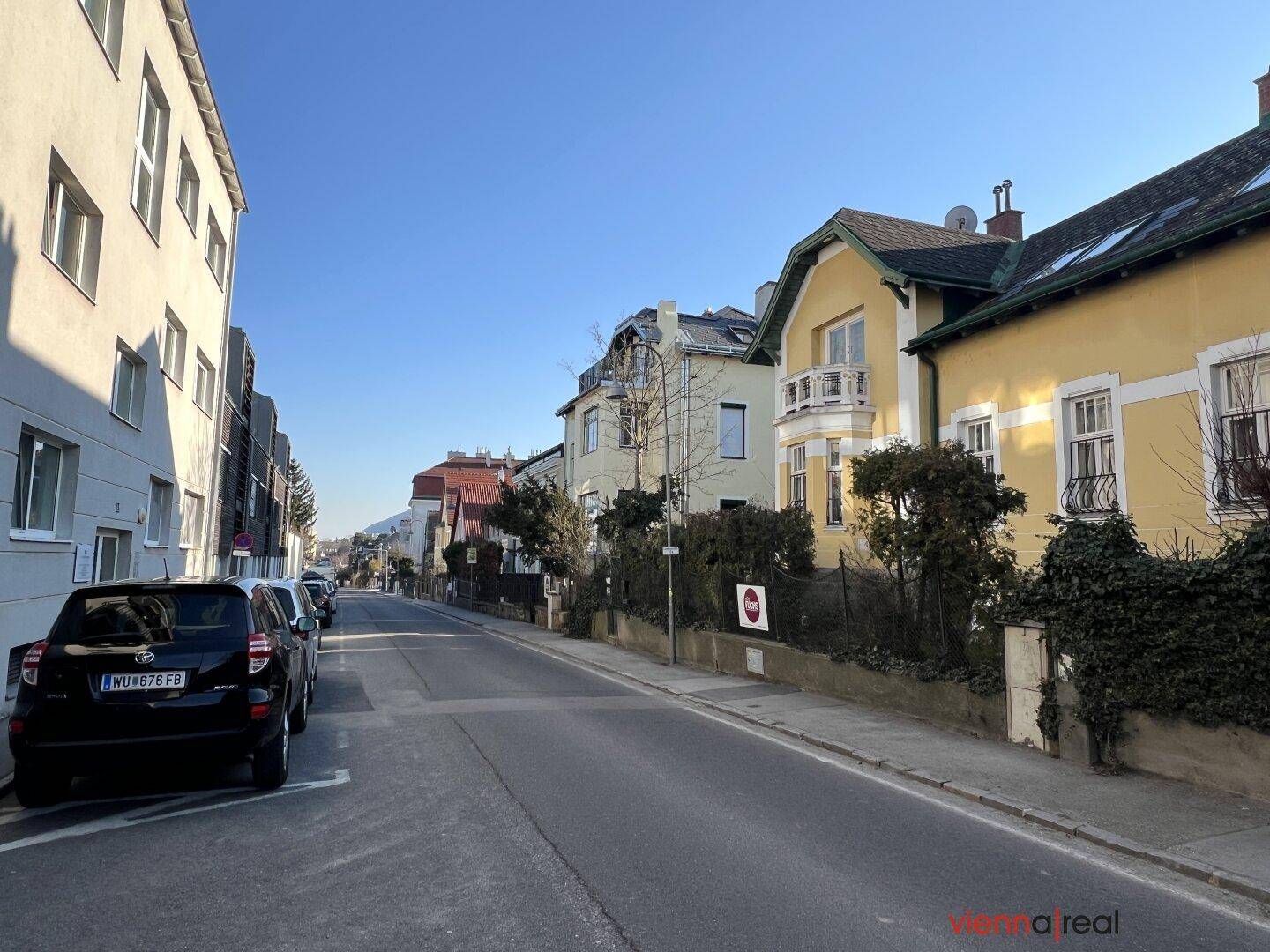 Wohnstraße mit verschiedenen Gebäuden und geparkten Autos unter blauem Himmel.