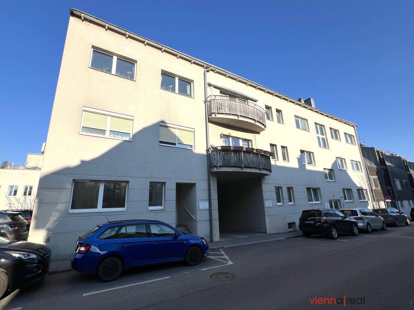 Modernes Mehrfamilienhaus mit Balkonen und einem Durchgang, umgeben von geparkten Autos.