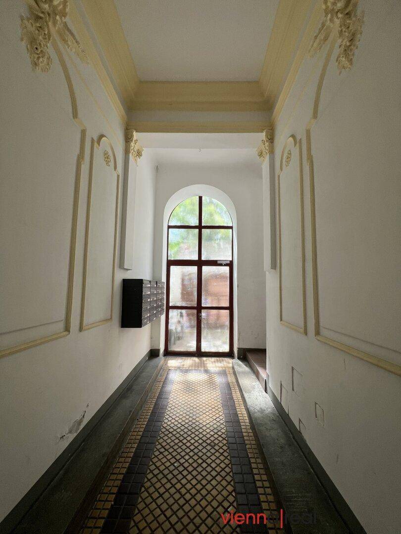 Langer Flur eines Altbaus mit originalen Fliesen, Stuckverzierungen und einem großen Bogenfenster am Ende.