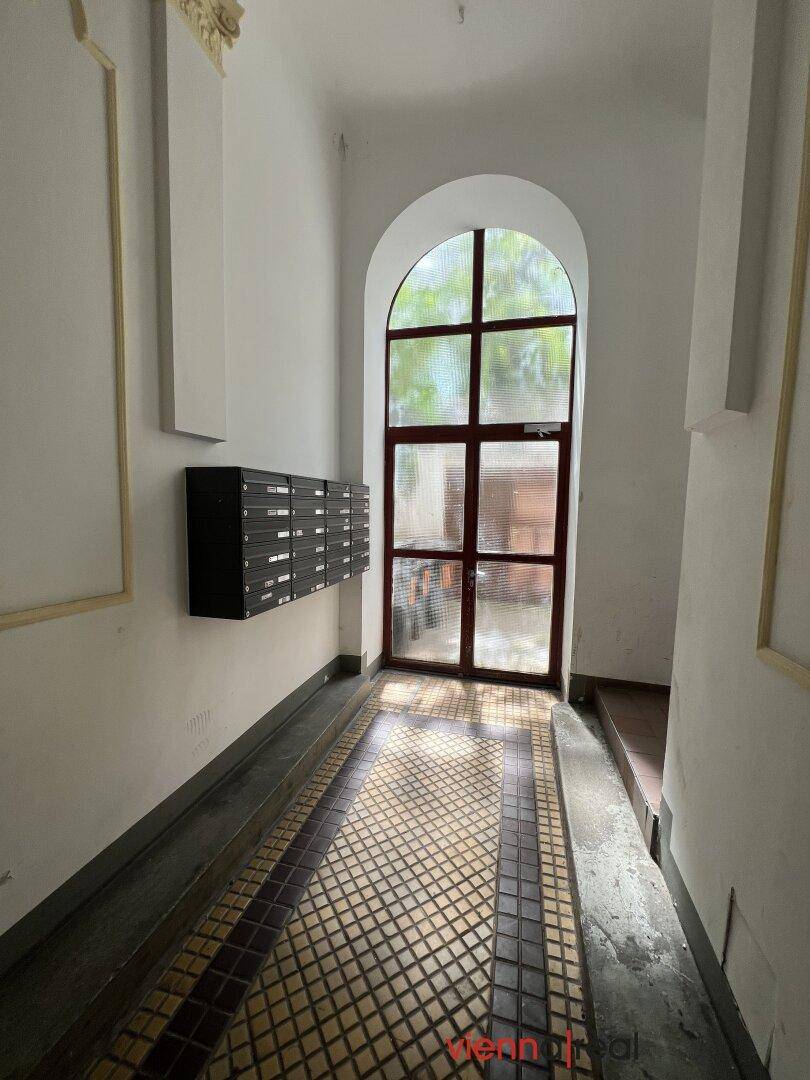 Heller Eingangsbereich mit historischen Fliesen, dekorativen Stuckelementen und einem großen Rundbogenfenster.