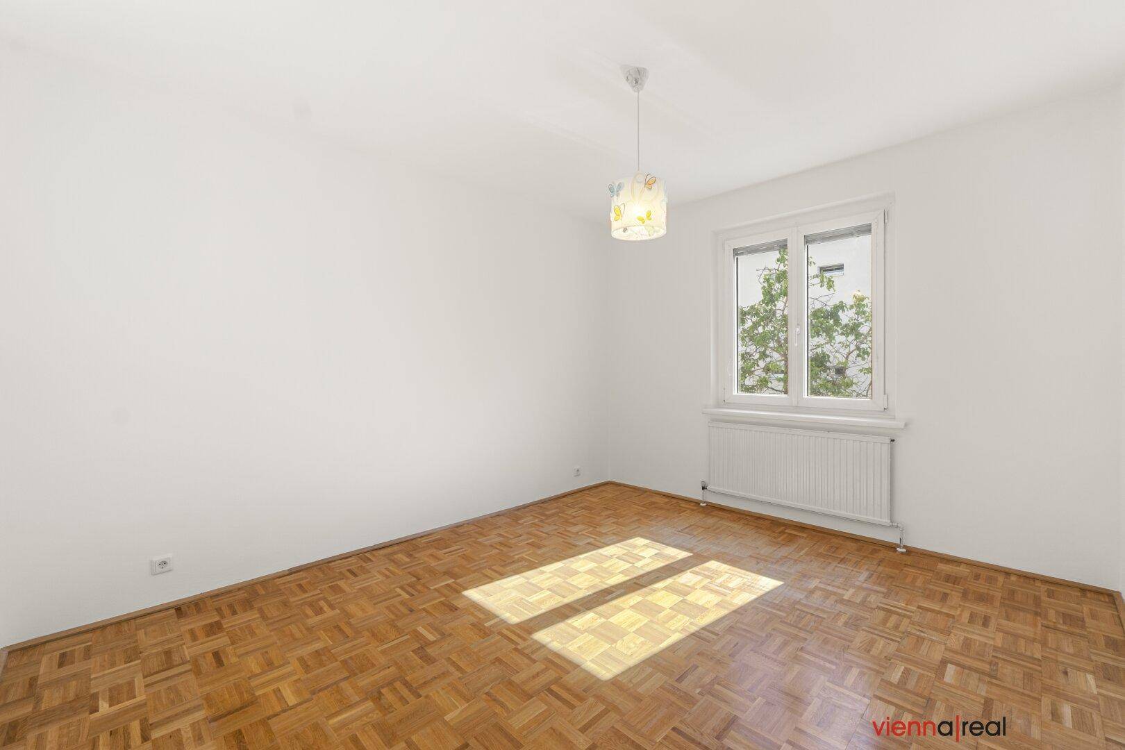 Helles Schlafzimmer mit Parkettboden und weiß gestrichenen Wänden, durchflutet von natürlichem Sonnenlicht.
