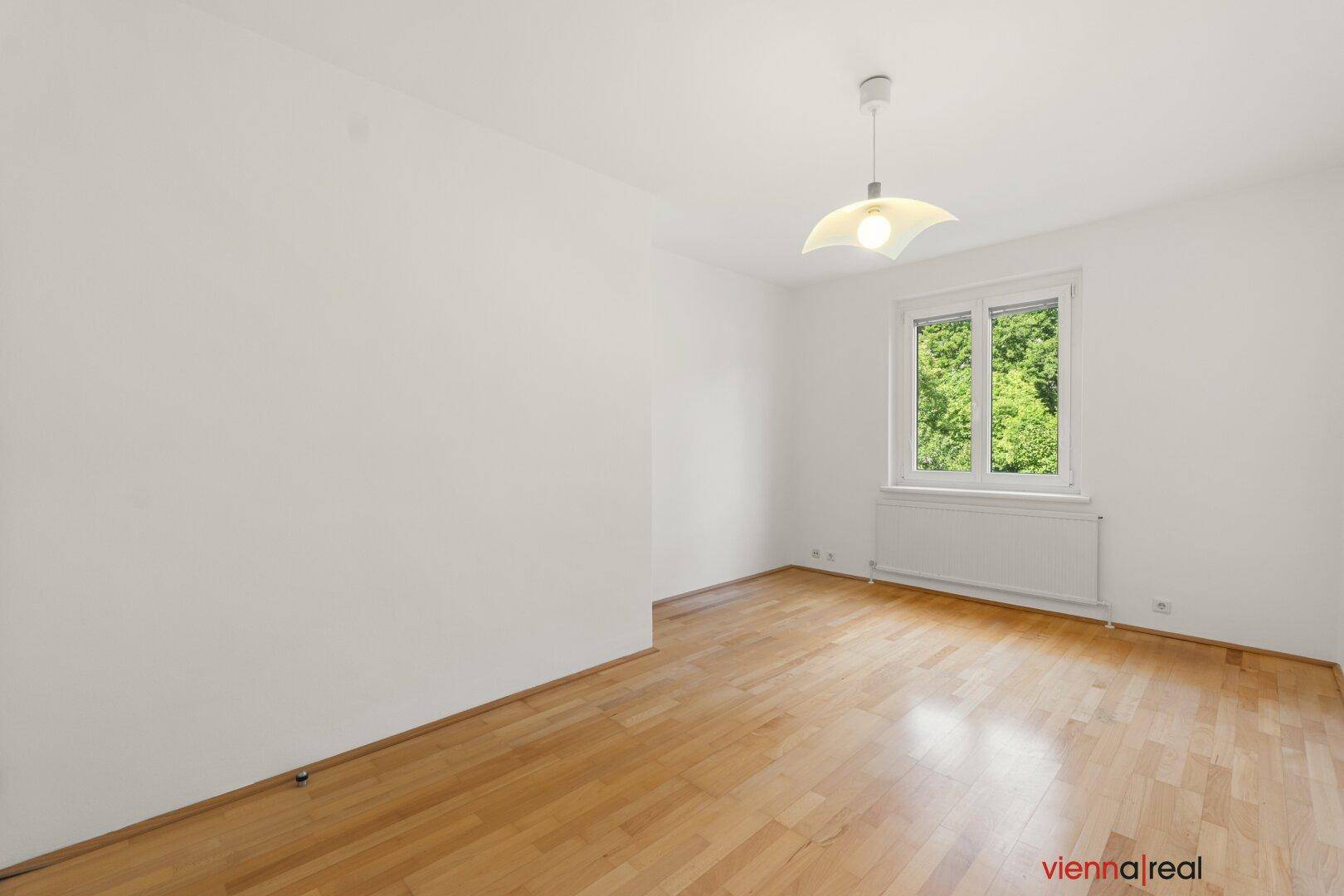 Geräumiges Zimmer mit hellem Parkettboden und Blick ins Grüne, ideal als Schlaf- oder Arbeitszimmer.