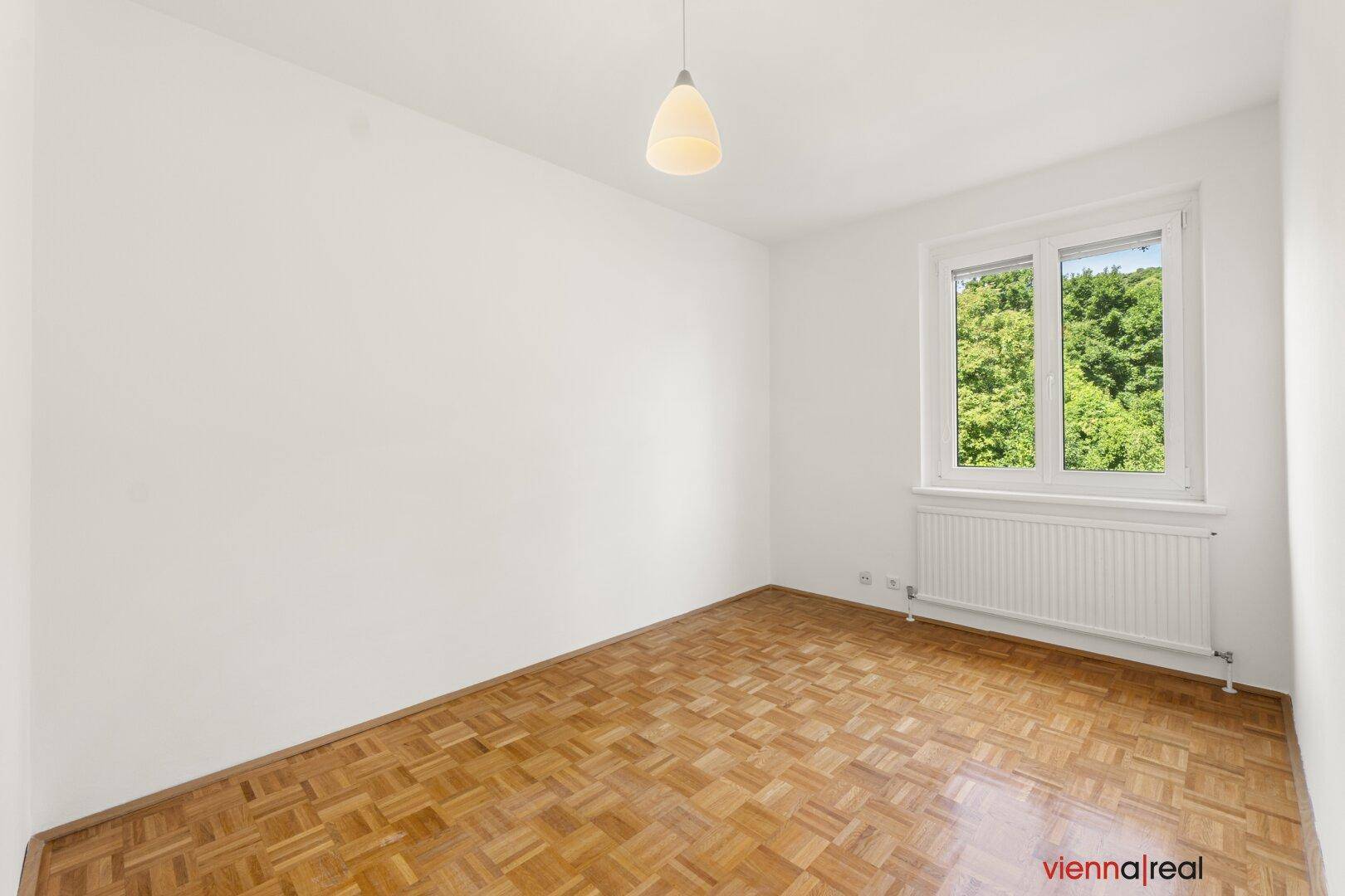 Freundliches Schlafzimmer mit klassischem Parkettboden und Fenster mit Ausblick auf grüne Bäume.