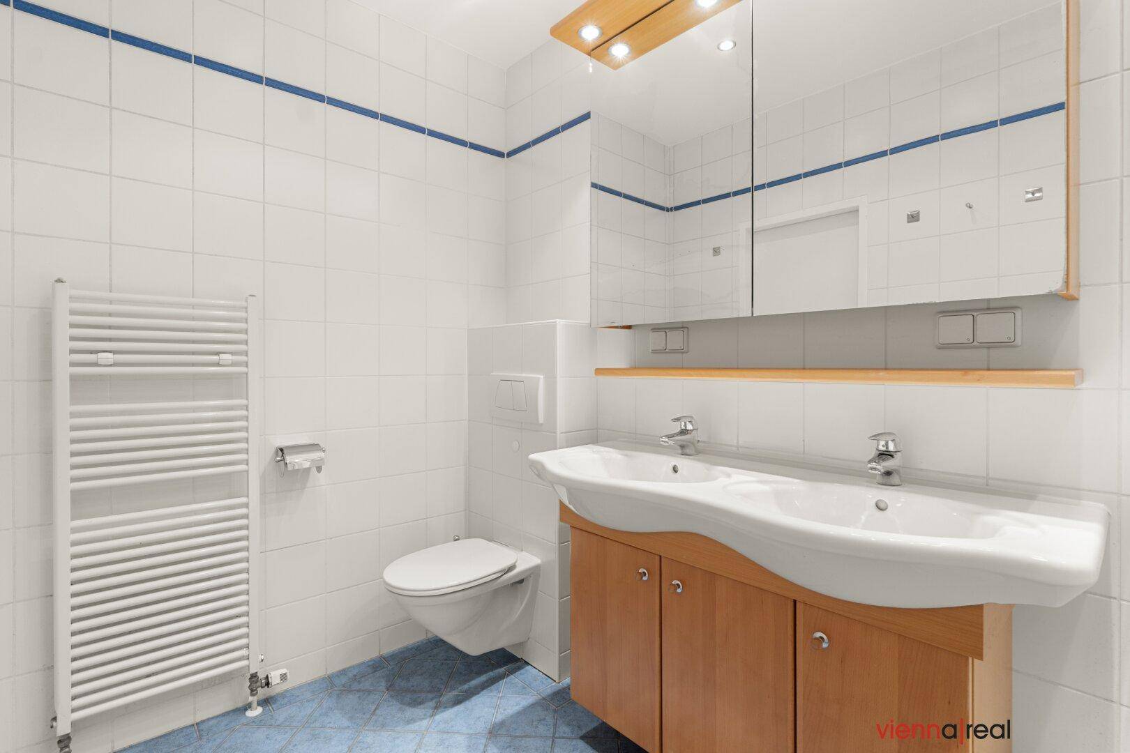 Badezimmer mit Wand-WC, Doppelwaschbecken und beheiztem Handtuchhalter, in hellen Fliesen gehalten.