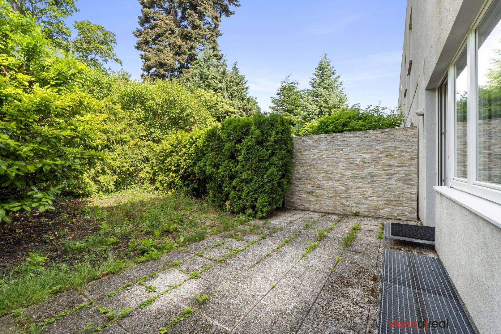 Gepflasterte Terrasse neben dem Gebäude, umgeben von Bäumen und Sträuchern, mit einer dekorativen Steinwand.
