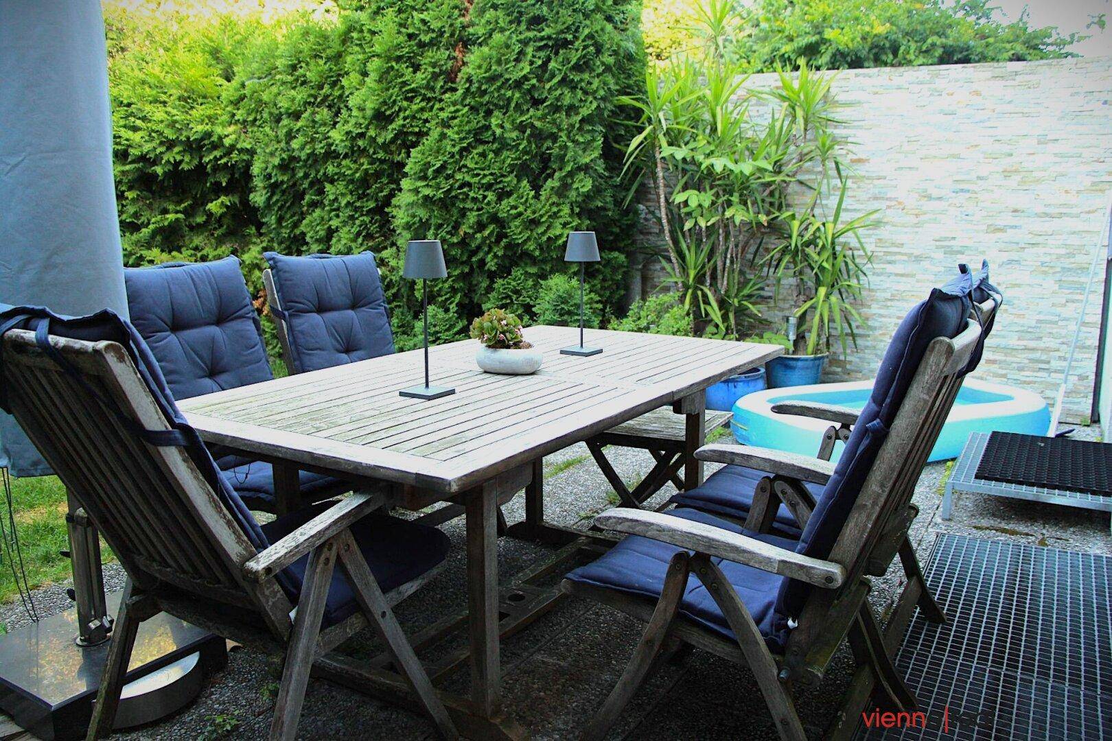 Gemütlicher Außenbereich mit Holztisch und Stühlen, umgeben von Pflanzen und einem kleinen Planschbecken.