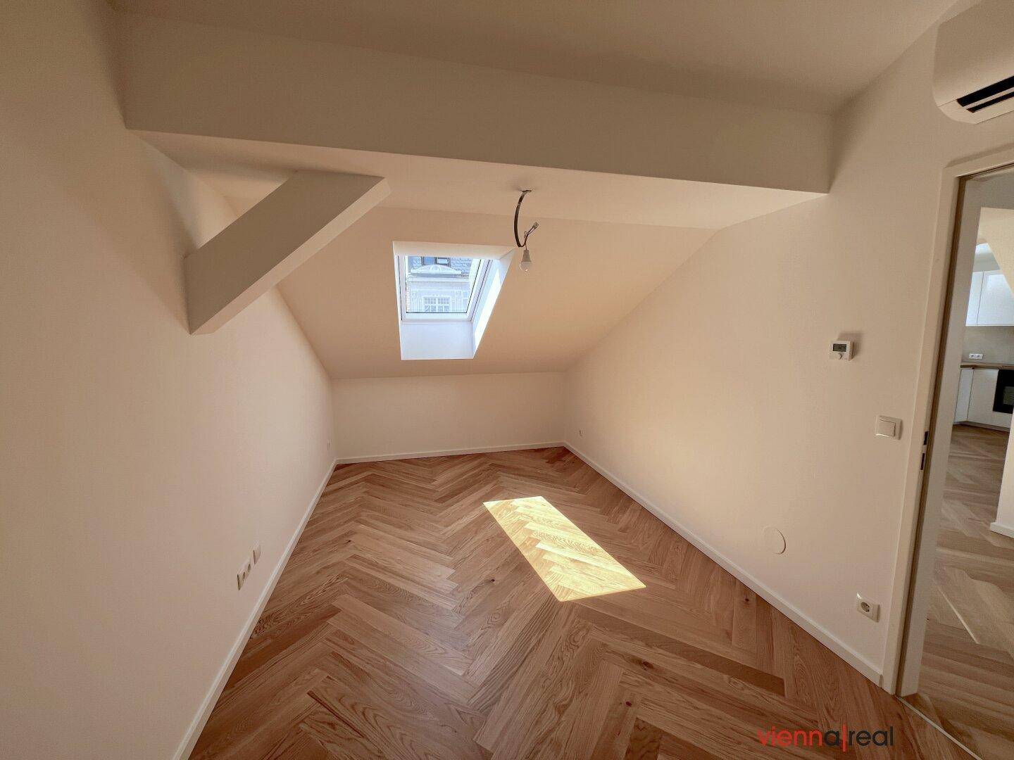 Heller Dachraum mit schrägen Decken, Dachfenster und elegantem Fischgrätparkettboden, ideal für Schlaf- oder Arbeitszimmer.