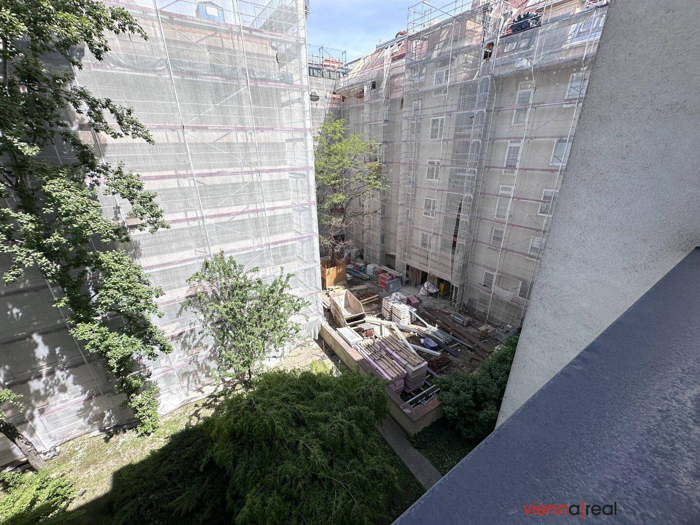 Blick von einem Balkon auf einen Innenhof mit Gebäuden unter Baugerüsten und Bäumen.