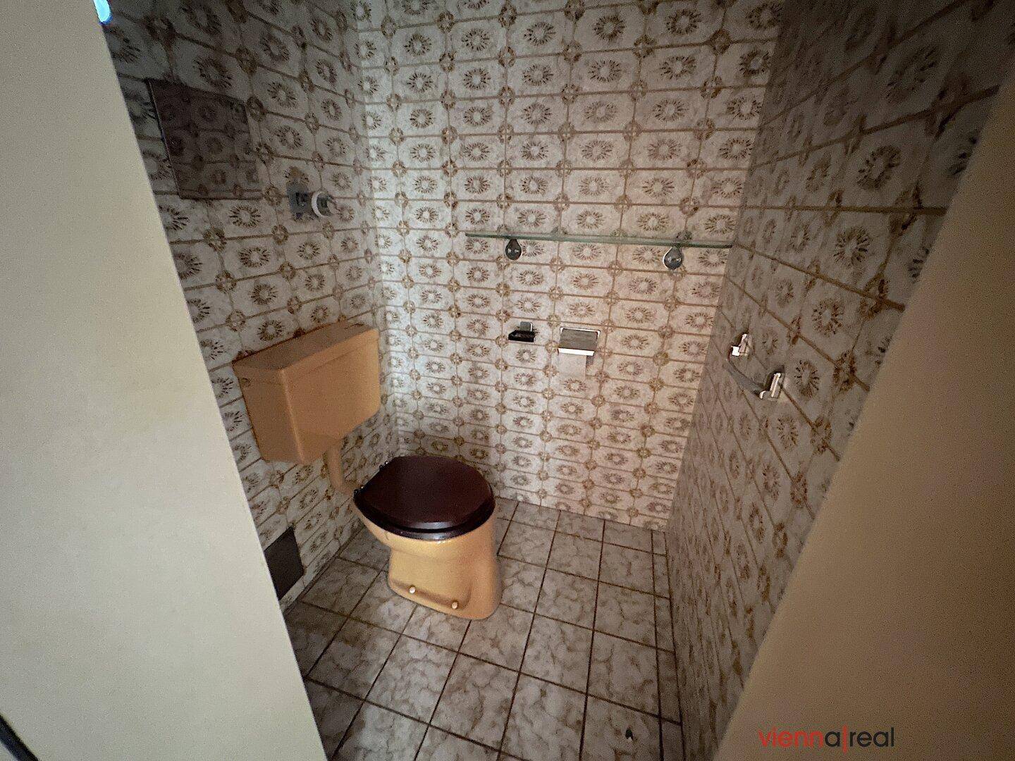 Veraltetes WC mit gemusterten Fliesen und einer Toilette im Originalzustand.