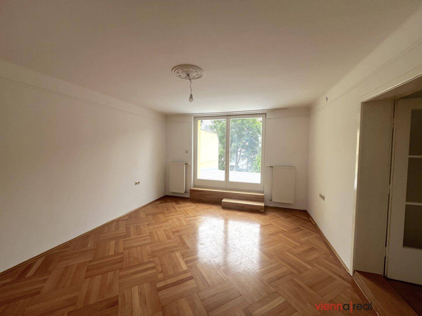 Geräumiges Zimmer mit klassischem Parkettboden und viel Tageslicht durch die große Balkontür.