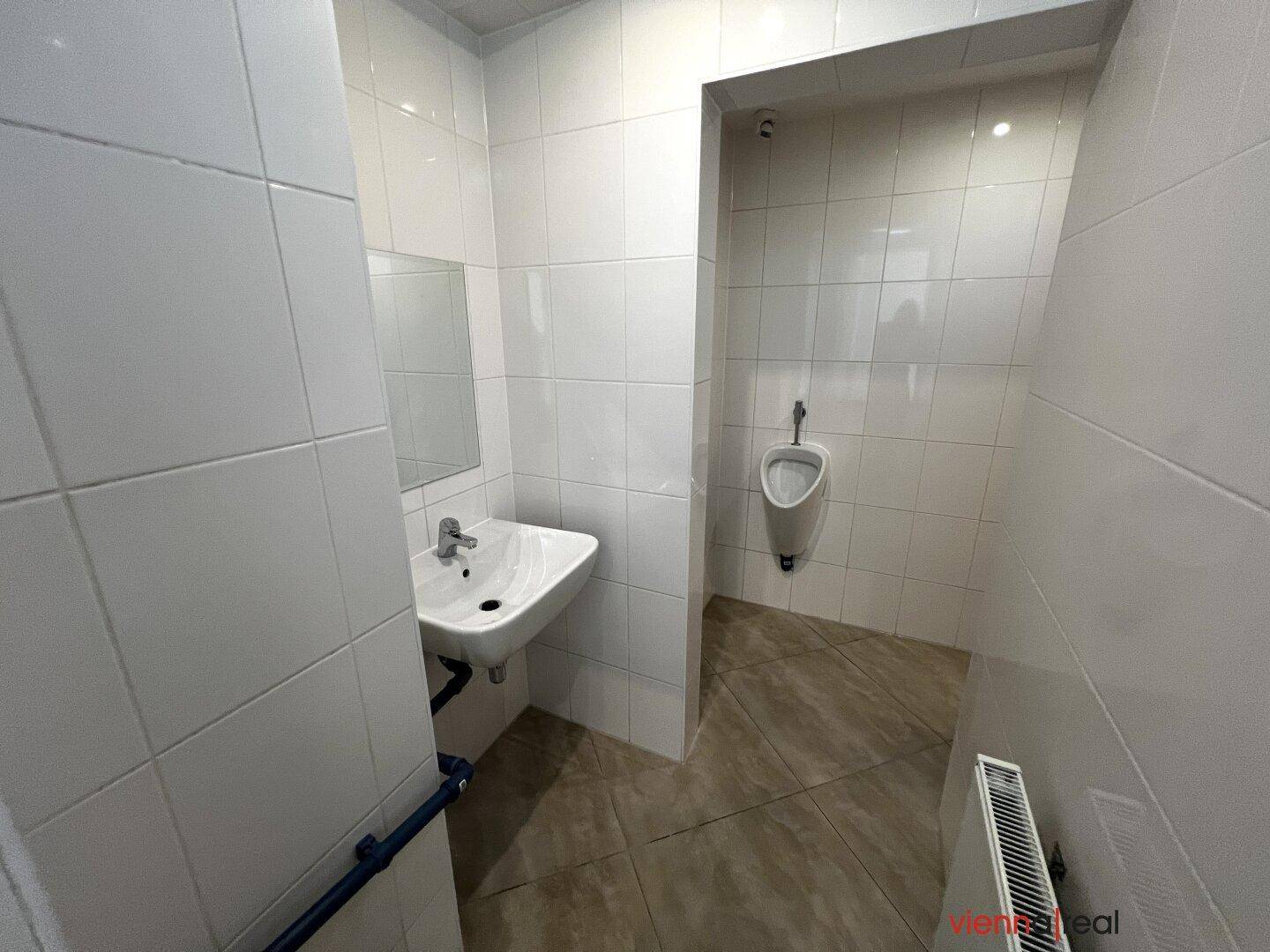 Gefliestes Badezimmer mit Waschbecken und einem Urinal im angrenzenden Bereich.