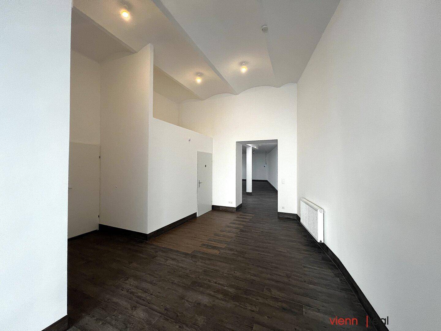 Langer, leerer Flur mit dunklem Holzboden und weißen Wänden, ausgestattet mit Deckenleuchten und Heizkörper.