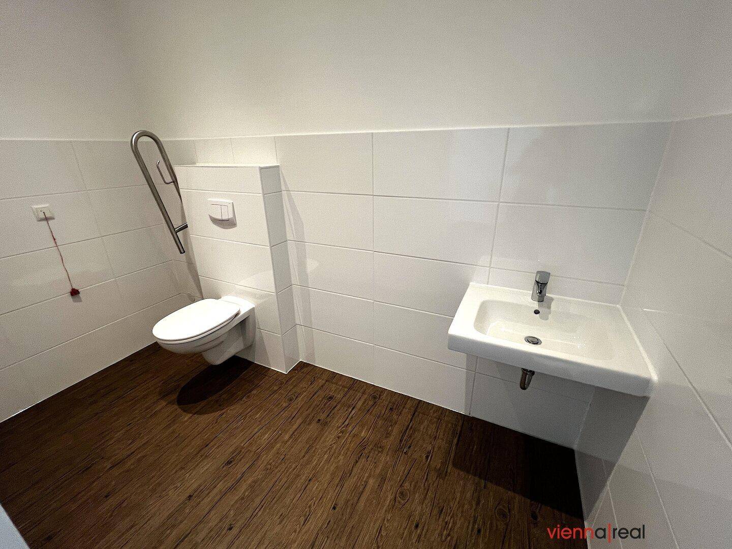 Barrierefreies Badezimmer mit Haltegriffen, Waschbecken und Holzoptik-Bodenfliesen.