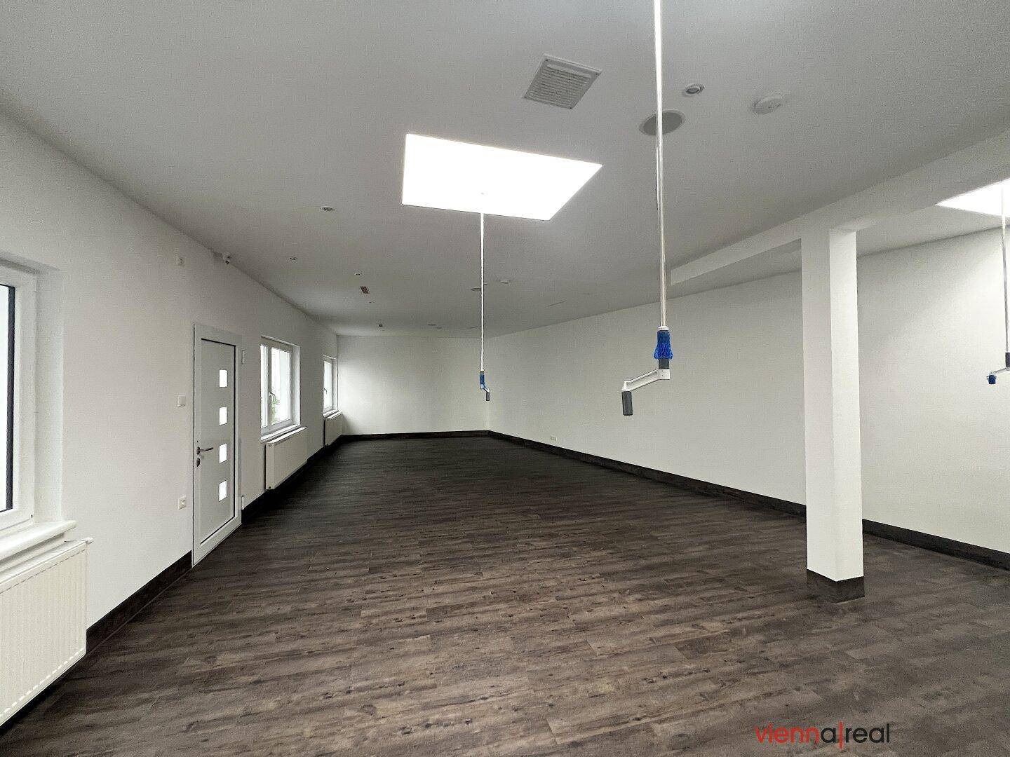 Weitläufiger, leerer Gewerberaum mit dunklem Holzboden, weißen Wänden und großen Fenstern sowie Oberlichtern.