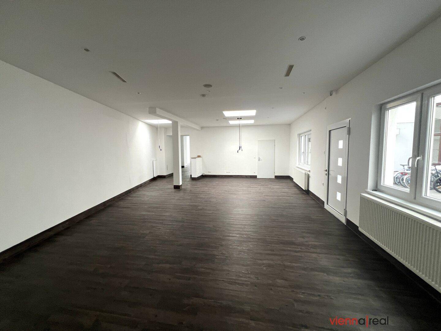 Geräumiger, leerer Gewerberaum mit dunklem Holzboden, weißen Wänden und mehreren Fenstern, bereit zur Nutzung.