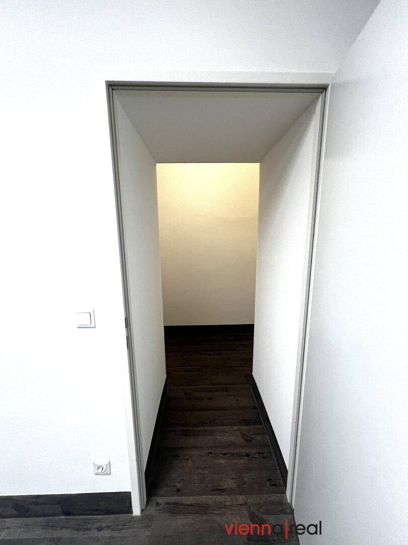 Schmaler Durchgang führt in einen weiteren leeren Bereich mit weißen Wänden und dunklem Holzboden.